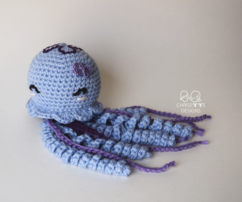 May include: A crocheted blue jellyfish with purple tentacles and a purple bow on its head. The jellyfish has embroidered eyes and a small flower on its head.