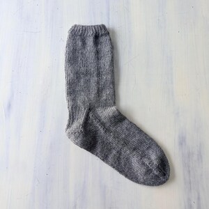 Puede incluir: Un calcetín gris tejido, tendido sobre una superficie blanca y azul claro. El calcetín está hecho de un hilo texturizado y tiene un puño acanalado. El calcetín es de color gris medio.