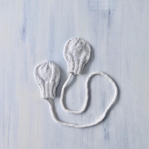 Op de afbeelding: Twee witte gebreide babywanten verbonden door een wit touwtje. De wanten hebben een bolvormige vorm met geribbelde manchetten. De achtergrond is een licht, getextureerd oppervlak.