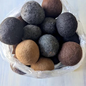 May include: A transparent bag containing a collection of felt balls in various sizes. The balls are in shades of brown, grey, and black. The felt balls have a soft, textured appearance.