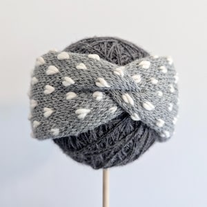 May include: A gray knit headband with white heart-shaped accents, displayed on a ball of dark gray yarn. The headband has a twisted design. The yarn ball is mounted on a wooden stick, against a white background.
