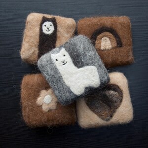 May include: Five square felted soaps in various shades of brown, grey and beige. Each soap features a different design: an alpaca, a rainbow, a flower, a heart, and another alpaca. The soaps are handmade.