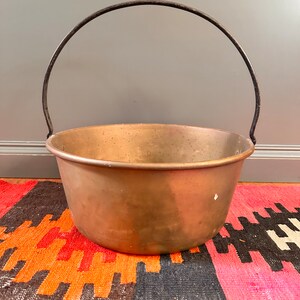 May include: A copper-colored metal cauldron with a curved black handle. The cauldron has a wide, rounded bowl shape. The background is a gray wall and a colorful rug with orange, black, and pink geometric patterns.