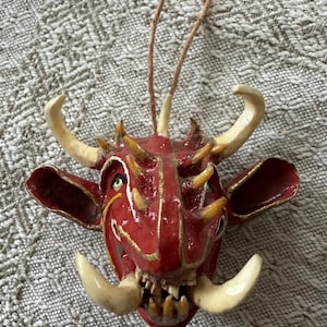 May include: A red, horned, and toothed ornament with gold accents. The ornament has a loop for hanging. The face has a menacing expression with large tusks and a detailed design. The ornament is likely for holiday or seasonal decor.