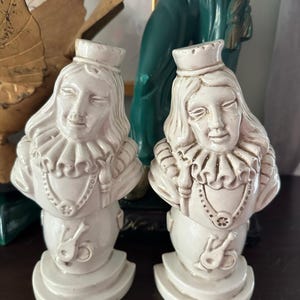 May include: Two off-white ceramic busts of a person with a ruffled collar, a crown, and a pendant necklace. The busts have a stylized design with a musical instrument on the base. The busts are displayed on a dark surface.