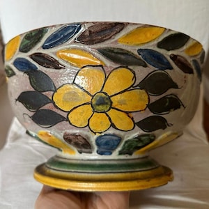 May include: A hand-painted ceramic bowl with a yellow flower design at the center. The rim features a leaf pattern in various colors, including yellow, blue, and brown. The bowl sits on a pedestal base with yellow and green stripes.