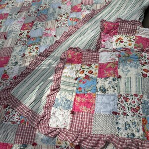 May include: A patchwork quilt with a variety of floral and striped patterns in shades of pink, blue, and white. The quilt has a ruffled edge with a red and white plaid design. The quilt is made of multiple fabric squares.