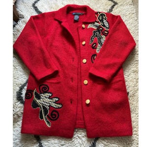 May include: A red wool coat with a notched collar and five round, gold-colored buttons. The coat features black and white checkered and black embroidered floral designs on the front and shoulders. The label reads "Diffusion".