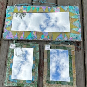 May include: Three mosaic mirrors with iridescent glass and a rectangular shape. The largest mirror has a yellow and iridescent border, while the smaller ones have green and blue borders. Each mirror reflects a cloudy sky and has a ribbon for hanging.