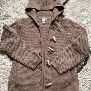 May include: A brown hooded sweater with a textured pattern and toggle closures. The sweater has a zipper closure and a hood. The sweater is made of a soft, knitted material. The sweater is a neutral color and can be worn by anyone.