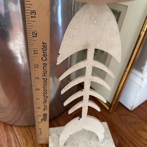 May include: A decorative, off-white fishbone-shaped candle holder. The candle holder is approximately 13 inches tall and rests on a square base. The fishbone design has a circular top for a candle. The candle holder has a distressed, textured finish.