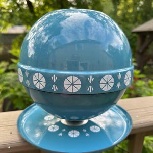 May include: A vintage teal-colored serving dish with a spherical top and a circular base. The dish features a white geometric pattern around the middle and on the base. The dish is made of ceramic or glass.