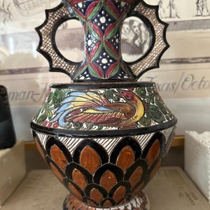 May include: A decorative ceramic vase with a detailed, colorful design. The vase features a bird motif, geometric patterns, and ornate handles. The color palette includes red, blue, green, and brown, creating a vibrant and artistic appearance.