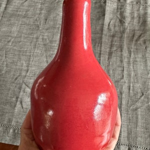 May include: A glossy, red ceramic vase with a rounded body and a narrow neck. The vase is held in a hand, showcasing its smooth, reflective surface. The background is a gray linen tablecloth.