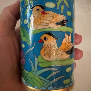 May include: Cylindrical blue cloisonné vase with gold trim, featuring two mandarin ducks on lily pads. The ducks have orange and black feathers, red beaks, and are set against a backdrop of aquatic plants.