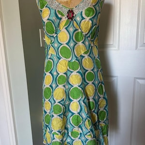 May include: A sleeveless dress with a teal, green, and yellow geometric pattern. The neckline is embellished with silver sequins and a red jewel. The dress is displayed on a vintage dress form.