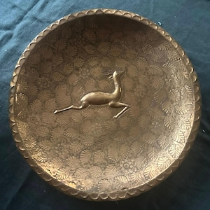 May include: A round, antique brass dish with a detailed floral and leaf pattern. The center features a raised image of a deer. The rim has a scalloped edge. The dish is approximately 8 inches in diameter.