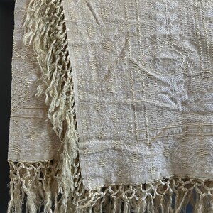 May include: Cream-colored textile with intricate woven patterns and long, knotted fringe. The fabric displays a detailed design with floral and geometric elements. The fringe hangs along the edges, adding texture and visual interest to the piece.