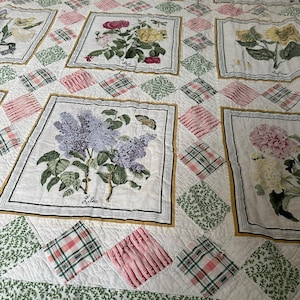 May include: A floral quilt featuring square panels with botanical illustrations of flowers like lilacs, roses, and sunflowers. The quilt has a white background with diamond-shaped patches in pink, green, and plaid patterns. The border is green with a yellow and white striped edge.