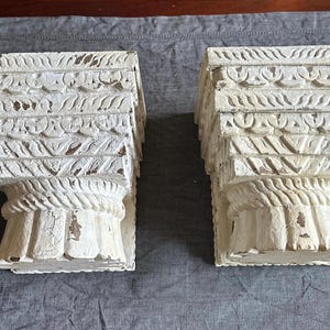 May include: Two distressed white decorative corbels with intricate carved details. The corbels have a layered design with a weathered, aged appearance. The bottom features a rope-like texture.
