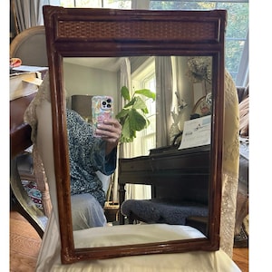 May include: A rectangular wall mirror featuring a dark brown bamboo frame with a woven rattan panel at the top. The mirror surface is clear and reflects the surrounding room, including a window, a piano, and a green potted plant.