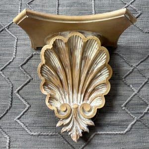 May include: A wall-mounted shelf featuring a decorative bracket shaped like a large scallop shell. The piece has a distressed finish with alternating tones of cream, white, and light brown, highlighting the carved ridges and scroll details.