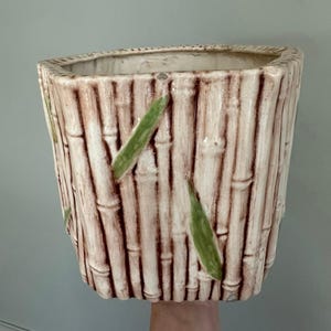 May include: A ceramic planter with a bamboo design. The planter is rectangular with a slightly trapezoidal shape. The background color is off-white with vertical brown lines to mimic bamboo stalks. Green leaves are painted on the front.