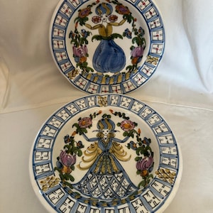 May include: Two decorative ceramic plates. Each plate features a central illustration of a woman in a blue dress, surrounded by floral and geometric patterns. The plates have a white base with blue and gold accents.