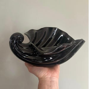 May include: A glossy black ceramic shell-shaped bowl. The bowl has a wavy, textured design with a curled accent on one side. The bowl is held in a hand against a neutral background. This decorative bowl is suitable for serving or display.