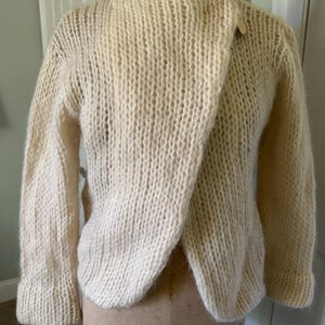 May include: Cream-colored, hand-knitted sweater with a unique wrap-style front closure. The sweater features long sleeves and a textured knit pattern. A single wooden button secures the wrap. The garment is displayed on a vintage dress form.