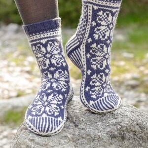May include: A pair of mid-calf knit socks featuring a dark blue and white fair isle pattern with floral motifs. The socks have a ribbed cuff, a reinforced heel, and a striped toe section. The design includes the text DROPS DESIGN www.garnstudio.com.