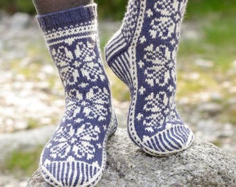 May include: A pair of mid-calf knit socks featuring a dark blue and white fair isle pattern with floral motifs. The socks have a ribbed cuff, a reinforced heel, and a striped toe section. The design includes the text DROPS DESIGN www.garnstudio.com.