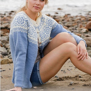 Puede incluir: Un cárdigan azul claro con un diseño estampado blanco y azul. El cárdigan tiene cierre de botones y lo lleva puesto una persona sentada en una playa de arena. La imagen incluye el texto "DROPS DESIGN. www.garnstudio.com".