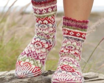 May include: A pair of colorful knitted socks with a geometric pattern. The socks are made with a variety of shades of pink, red, green, and white yarn. The socks are shown on a person's legs, with the feet resting on a rock.