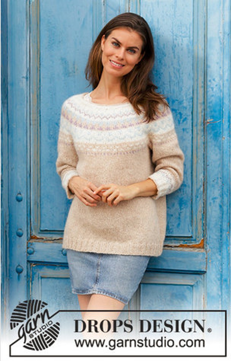 Puede incluir: Un su&eacute;ter beige con un patr&oacute;n geom&eacute;trico colorido alrededor del cuello. El su&eacute;ter lo lleva una persona de pie frente a una puerta de madera azul. El texto "DROPS DESIGN." y "www.garnstudio.com" es visible en la parte inferior de la imagen.