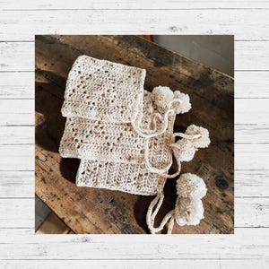 Pode incluir: Um conjunto de três gorros de croché na cor creme com um padrão salpicado em todo o fio. Cada gorro tem uma coroa quadrada, uma aba estruturada e laços longos terminados com grandes pompons felpudos.