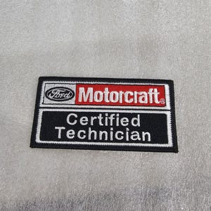 May include: A rectangular embroidered patch featuring a black border and white trim. The top section displays the Ford logo and the word Motorcraft in white text on a red background. The bottom section contains the text Certified Technician in white.