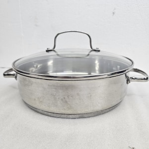 May include: A round stainless steel cooking pot with a polished finish, featuring two side handles and a clear glass lid with a central metal handle. The pot has a wide, shallow design with a flat base and a smooth, reflective metallic surface.