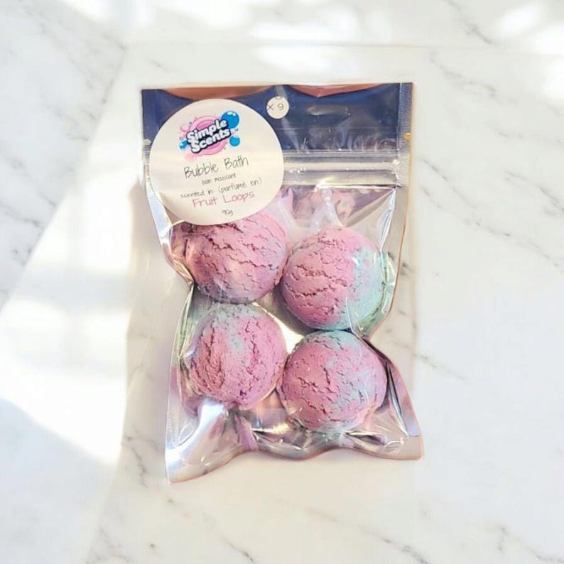 May include: A clear plastic bag containing four spherical bubble bath bombs. The bath bombs are a mix of pink and blue, resembling scoops of ice cream. The bag has a label that reads "Simple Scents Bubble Bath" and "Fruit Loops".