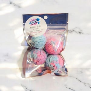 May include: A clear plastic bag containing four bubble bath bombs. The bath bombs are a mix of pink and blue colors. The bag has a white label with the text "Simple Scents Bubble Bath" and "Monkey Farts".