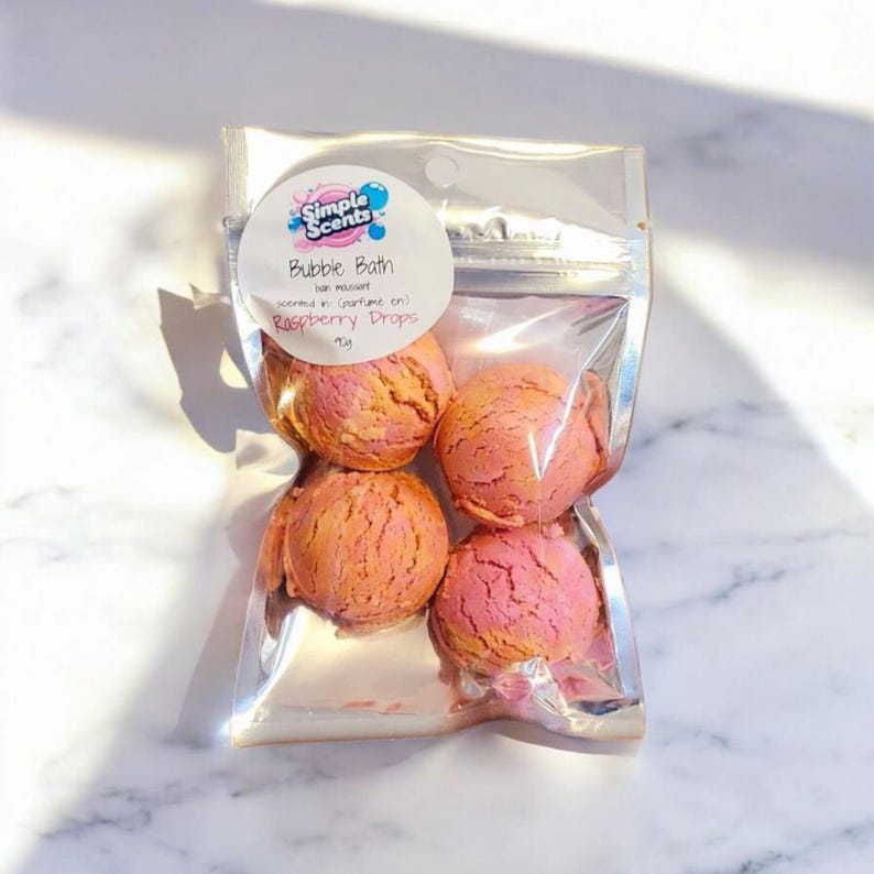 May include: A clear plastic bag containing four raspberry-scented bubble bath spheres. The spheres are pink and orange, with a textured surface. The bag has a label that reads "Simple Scents Bubble Bath Raspberry Drops".