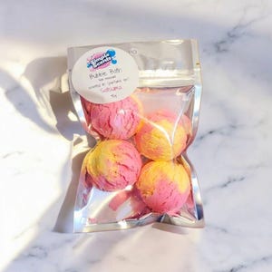 May include: A clear plastic bag containing four bubble bath spheres. The spheres are a mix of pink and yellow, with a textured surface. The bag has a white label that reads "Simple Scents Bubble Bath" and "Satsuma".