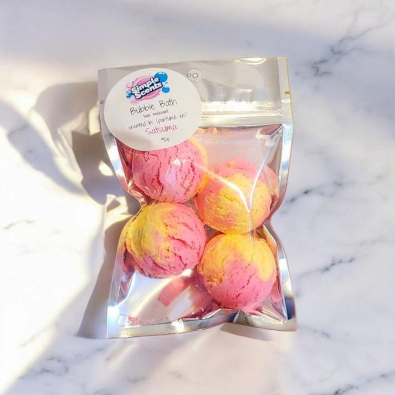 May include: A clear plastic bag containing four bubble bath spheres. The spheres are a mix of pink and yellow, with a textured surface. The bag has a white label that reads "Simple Scents Bubble Bath" and "Satsuma".