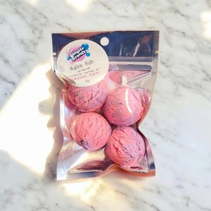 May include: A clear plastic bag containing five pink and purple bubble bath bombs. The bag has a label that reads "Simple Scents Bubble Bath" and "Unicorn Farts". The bath bombs are spherical and have a textured surface.