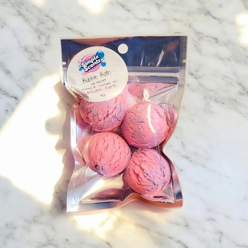 May include: A clear plastic bag containing five pink and purple bubble bath bombs. The bag has a label that reads "Simple Scents Bubble Bath" and "Unicorn Farts". The bath bombs are spherical and have a textured surface.