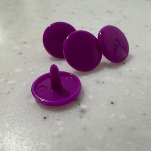 May include: A collection of four purple snap fasteners. One fastener is open, revealing the post, while the others are closed. The fasteners are circular and made of a smooth, glossy material. They are scattered on a light-colored surface.