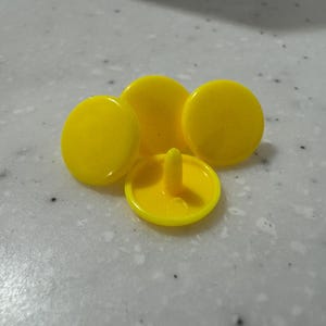 Peut inclure: Ensemble de cinq boutons-pression jaunes. Quatre sont circulaires avec une surface lisse et brillante, et un a une tige et une zone encastrée. Les boutons sont disposés sur une surface grise clair mouchetée.