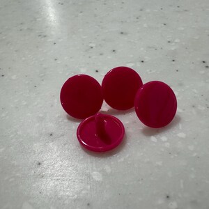 Può includere: Quattro bottoni rosa acceso, rotondi e lucidi. Un bottone è capovolto, mostrando il gambo. I bottoni sono sparsi su una superficie chiara e macchiettata. Sembrano essere fatti di plastica o un materiale simile.