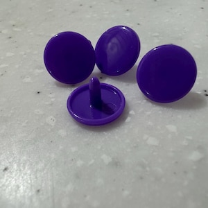 Pode incluir: Quatro fechos de pressão roxos. Três são planos e redondos, enquanto o quarto tem um pino na parte inferior. Os fechos são feitos de um material liso e sólido e têm um tom roxo vibrante.