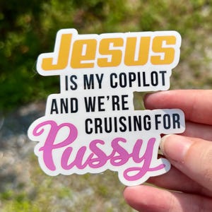 May include: A white sticker with the text "Jesus is my copilot and we're cruising for pussy." The word "Jesus" is in yellow with an orange outline. The rest of the text is in black, with the word "pussy" in pink.